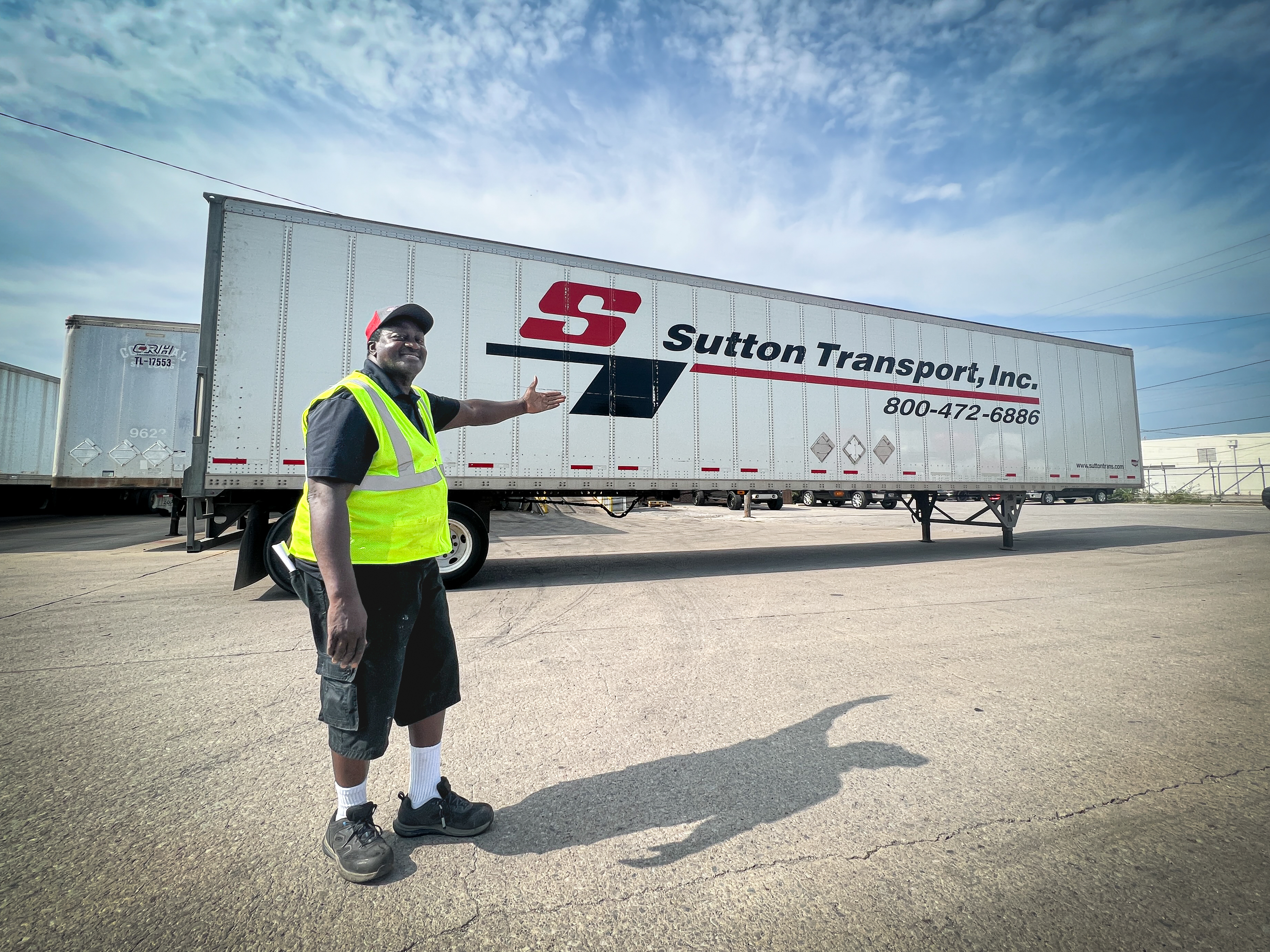 A cheerful Sutton Transport truck driver proudly points to a company truck, expressing happiness and pride in their work.