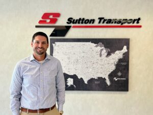 Cliff Sutton, President of Sutton Transport, standing proudly in front of the company sign with a map of America in the background.
